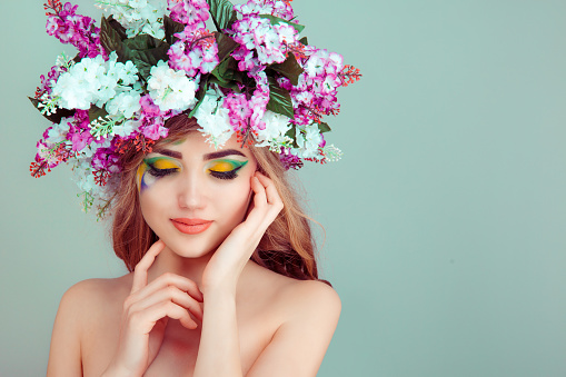 Beautiful young woman smiling model with flowers on head floral headband hairstyle makeup yellow and green eyeshadow closed eyes isolated on light green background. Spring woman concept