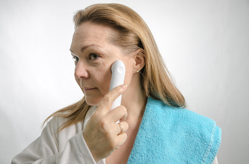 Woman in bathrobe using an ultrasound skin care device on cheek