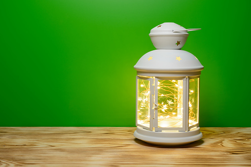 A lantern with a glowing garland in on the table, green background