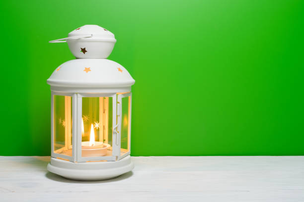 A lantern with a glowing candle in on the wooden table, green background