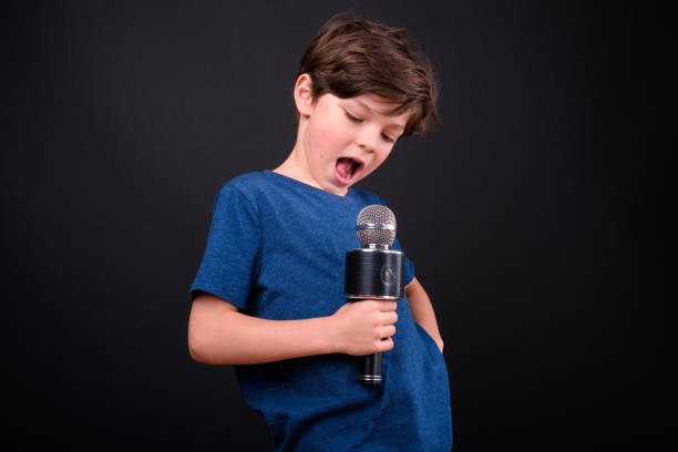 istock Portrait Of Cute Boy Singer Using Microphone Against Black Background 929473402