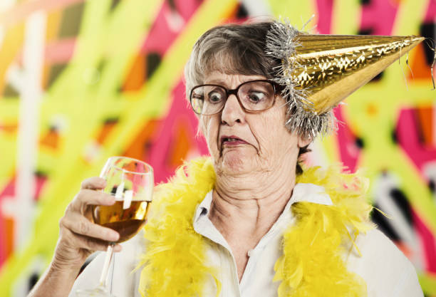 Old woman in party hat looks at wine glass, horrified An old woman wearing a party hat and feather boa holds up a wine glass, looking down at it with a disgusted and somewhat drunken expression on her face. ugly old women stock pictures, royalty-free photos & images