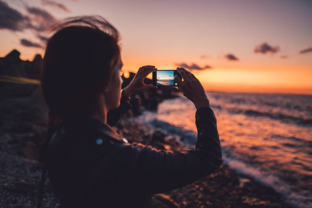 mujer en la playa de fotografiar la puesta de sol - dispositivo de información móvil fotos fotografías e imágenes de stock