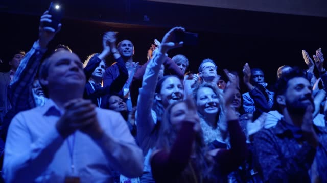 Enthusiastic audience giving standing ovation, clapping in dark conference audience
