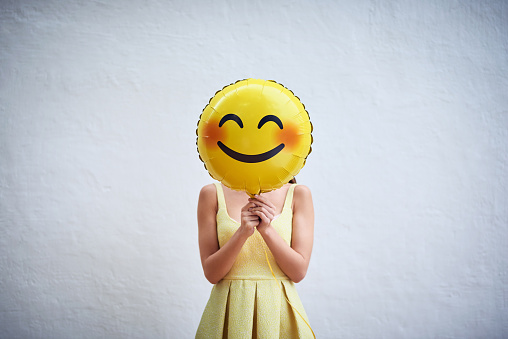 Studio shot of a young woman holding a balloon with a smiley in front of her face