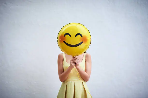 Studio shot of a young woman holding a balloon with a smiley in front of her face