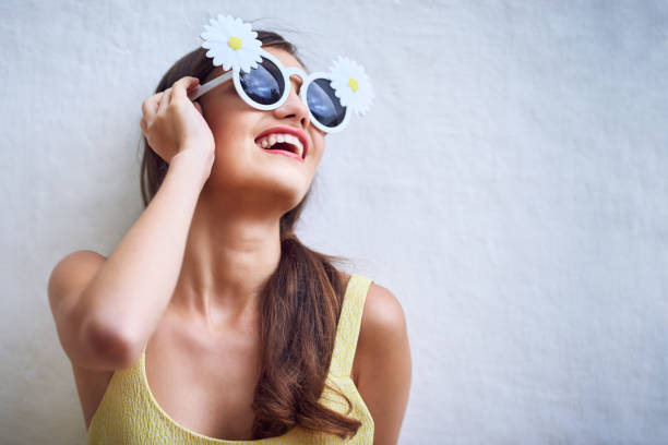 Little blonde-haired girl with green sunglasses holding a yellow flower while posing with a curious expression in front of a textured brick and cement wall. Image conveying freshness, spontaneity and contrast between urban and natural.