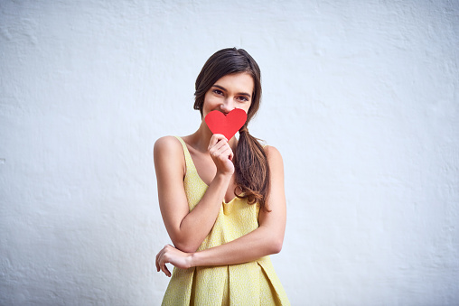 Studio shot of a cheerful young woman holding a heart shaped piece of paper in her hands while standing against a grey background