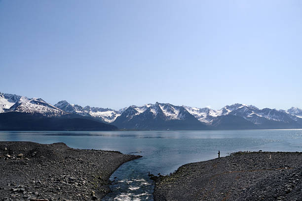 A lone fisherman silhouetted against Resurrection Bay in Seward, Alaska. He is fishing at a rill flowing into the bay. the Chugach Mountains are in the background and the sky is cloudless and blue.