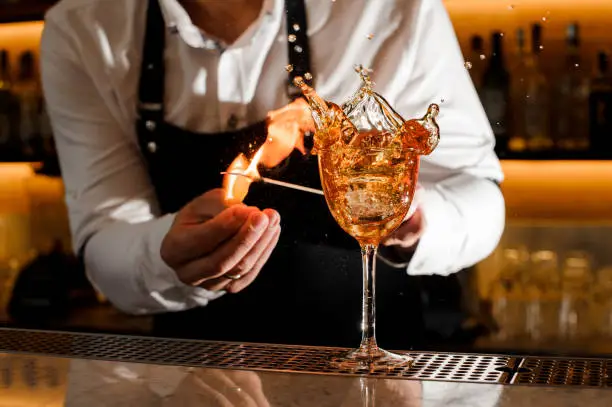 Barman making a fresh burning cocktail with fire on the bar counter against the shelves with bottles