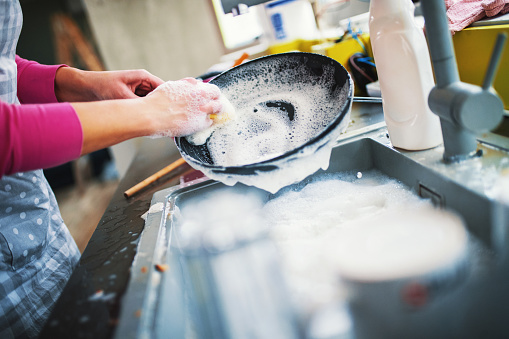 Closeup of unrecognizable woman doing dishes with lots of dirty plates and pots waiting aside. She's cleaning a black non stick cooking pan.