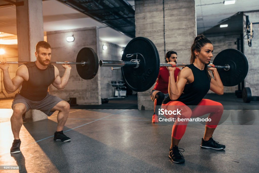 No pain, no gain Shot of two young men and a woman lifting weights at the gym Squatting Position Stock Photo