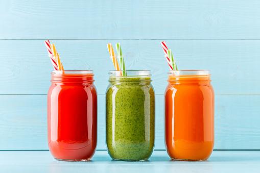 Orange/green/red colored smoothies / juice in a jar on a blue background.