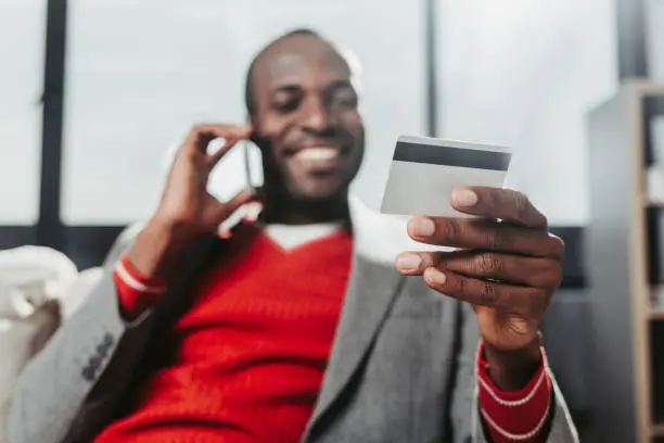 Low angle portrait of delighted man chatting by mobile phone at home. Focus on hand holding credit card