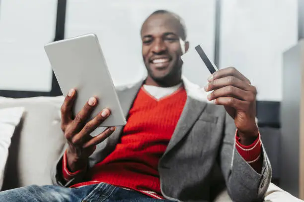 Low angle portrait of happy african man sitting on couch and holding gadget and payment card. Focus on arms with tab and plastic