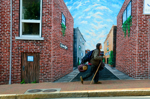 Asheville, North Carolina, USA - April 28, 2012: A young man on a city sidewalk, carrying bags and using a walking stick, walks past a mural painted on a wall. Asheville is known for its art scene and alternative lifestyles.