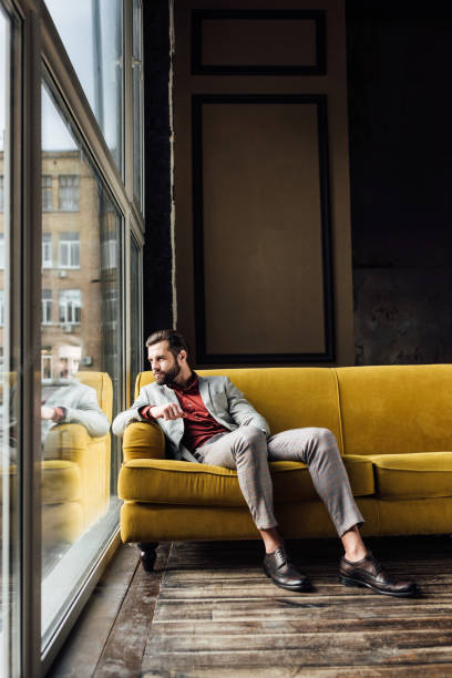 istock handsome fashionable man sitting on yellow sofa and looking at window 929803266