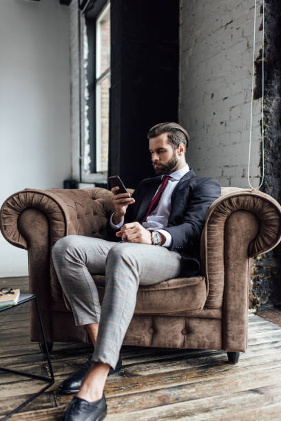 istock confident businessman texting sms on smartphone while sitting in armchair in loft 929818760