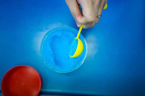 Hands holding spoon of blue chemical, Copper Sulphate Pentahydrate, against blue table for junior scientific lab test and preparation.