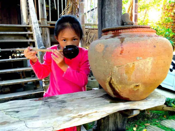 Little girl drinking water with dipper or scoop coconutshell ladle from Old clay jar and old wooden house background.