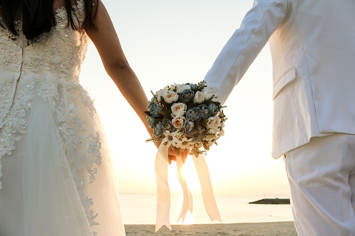 Young and smiling newlyweds are walking along the seashore, holding hands and looking at each other against the background of a beautiful seascape