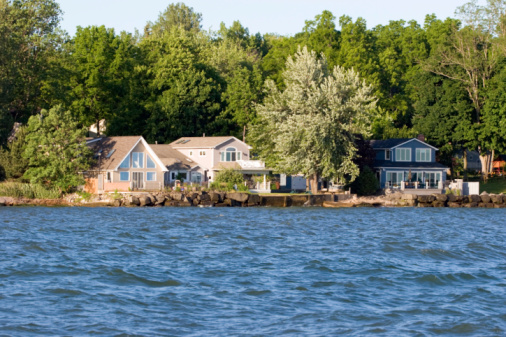 Wide view of two lake houses on Lake Ontario in New York, on of the Great Lakes with blue water in the foreground and gree trees in the background.  Though there are waves on the water, it is relatively calm for the Great Lakes.  There are no people in the image and ample copy space for text