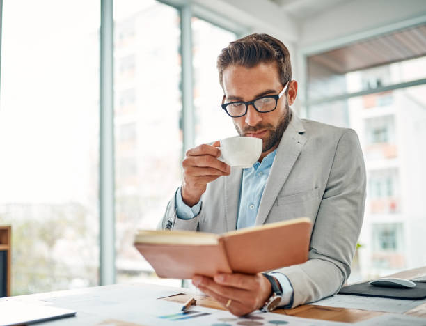 He's a well-read entrepreneur Shot of a handsome young businessman drinking coffee while reading a book in an office Business Books stock pictures, royalty-free photos & images