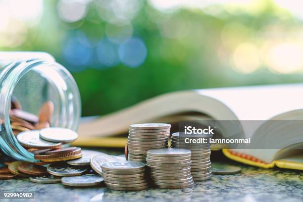 Pile Of Money Coins In And Outside The Glass Jar On Blurred Book And Natural Green Background For Financial And Education Concept Stock Photo - Download Image Now