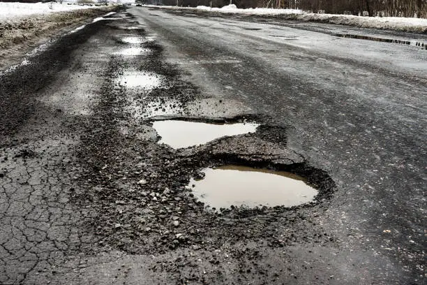 old highway with holes and snow. Landscape road in the potholes in cloudy winter weather. The concept of the absence of timely repair of the highway.