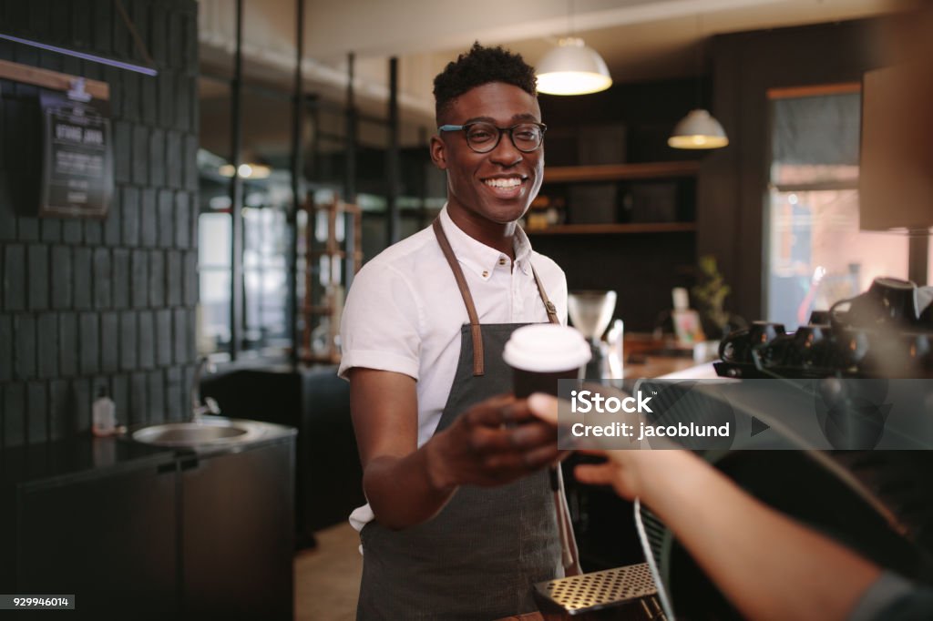 Barista serving customers inside a coffee shop Coffee shop owner handing over a sealed coffee cup to a customer. Man serving customer with a smile at a coffee shop. Barista Stock Photo