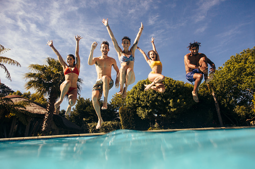 Group of friends jumping in swimming pool with their hands raised. Men and women jumping in pool and having fun.