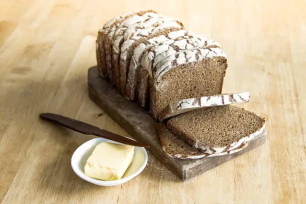 Sliced loaf of dark rye bread on a wooden board, with butter beside it