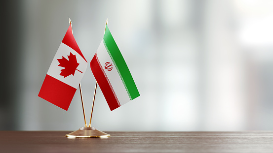 Canadian and Iranian flag pair on desk over defocused background. Horizontal composition with copy space and selective focus.