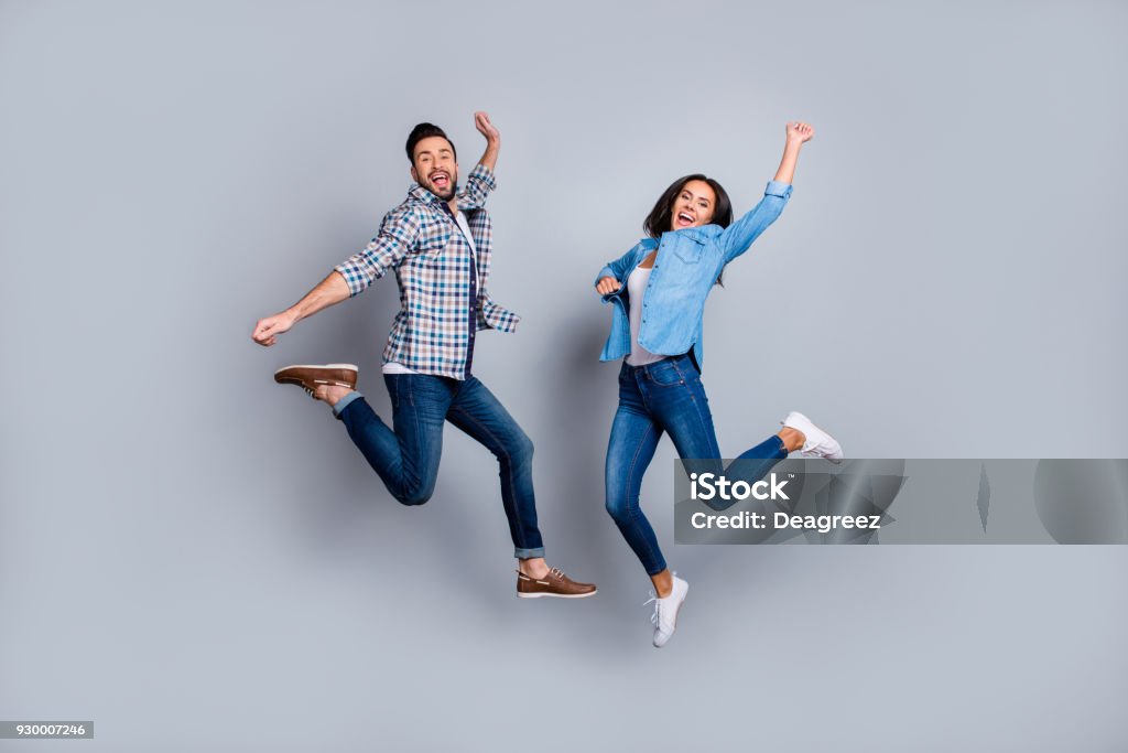 Vs retrato de longitud completa de de pareja atractiva, juguetona, alegre, cómica en ropa casual, jeans, camisas saltando sobre fondo gris - Foto de stock de Saltar - Actividad física libre de derechos