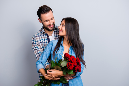 Portrait of cheerful lovely cute couple with beaming smiles hugging and looking to each other, woman hold a bouquet of red roses, family celebrating valentine day over grey background