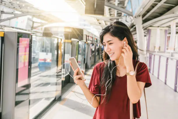 Photo of Asian woman passenger with casual suit using and listening the song via smart mobile phone in the BTS Skytrain rails or MRT subway for travel in the big city, lifestyle and transportation concept