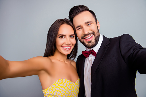 Lovely, cute, smiling, attractive, sexy couple, husband and wife in elegant outfit, tux, dress with beaming smiles making selfie together in two hands over grey background, 14 february