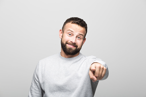 Portrait of happy bearded young man pointing at camera. Studio shot, grey background.