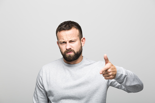 Portrait of angry bearded young man pointing at camera with index finger. Studio shot, grey background.