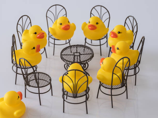 istock Group of eight yellow rubber ducks sitting on chairs facing each other around a table, with one empty chair and a duck rushing towards the chair as if late for a meeting, scene set on artificial grass. 930034492