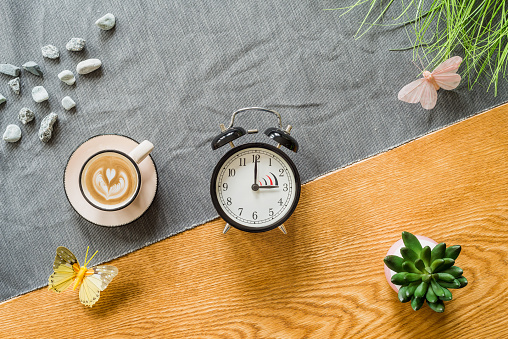 Change to daylight saving time photo of alarm clock with coffee from above as flatlay with wooden background in summer
