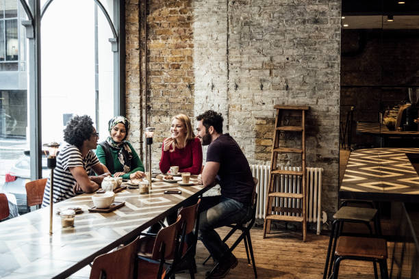 Four friends sitting at long table in cafe by window talking Multi racial group of people talking and smiling in restaurant, man with afro hairstyle talking, man and two women litening hijab photos stock pictures, royalty-free photos & images