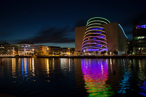 The Dublin Convention Centre on North Wall Quay lit up in rainbow colors, with a rainbow reflection in the Liffey river.