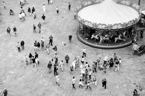 Black and white depiction of people walking through a town square and enjoying a carousel.