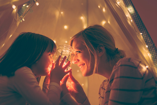 Mother and daughter sitting in a tent, holding a jar with christmas lights and telling stories. Focus on the mother