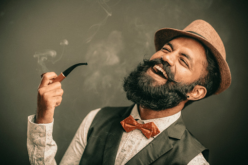 Laughing bearded young man with hat and smoker on dark.