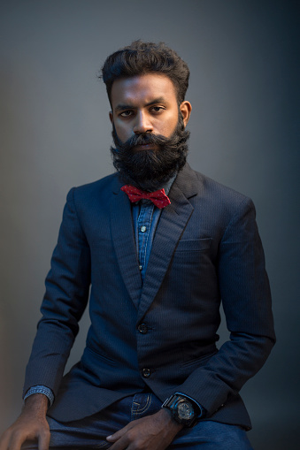 Portrait of young man posing with beard in suit on gray background.