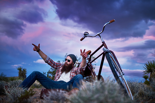 bearded traveler with suitcase and bike in the desert shouting with arms up