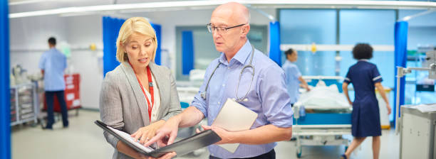hospital business a female businesswoman chats to a senior doctor on a busy hospital ward rolled up sleeves stock pictures, royalty-free photos & images
