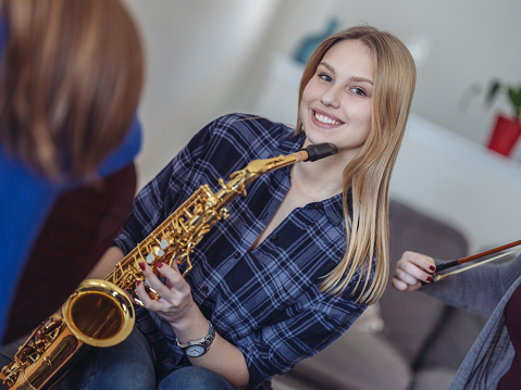 Group of 3 teenage girls having music party at home. Playing some great tunes on violin, saxophone and guitar. Dressed in casual clothing. Focus on the young woman in the background playing saxophone.